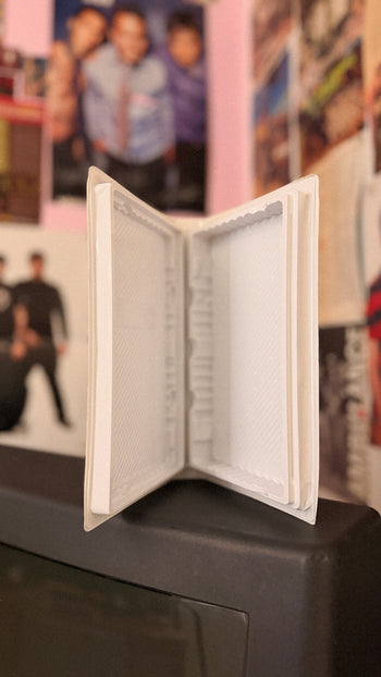 A white, open book-shaped frame or organizer standing on a dark surface, with a blurred indoor background featuring posters and photos.