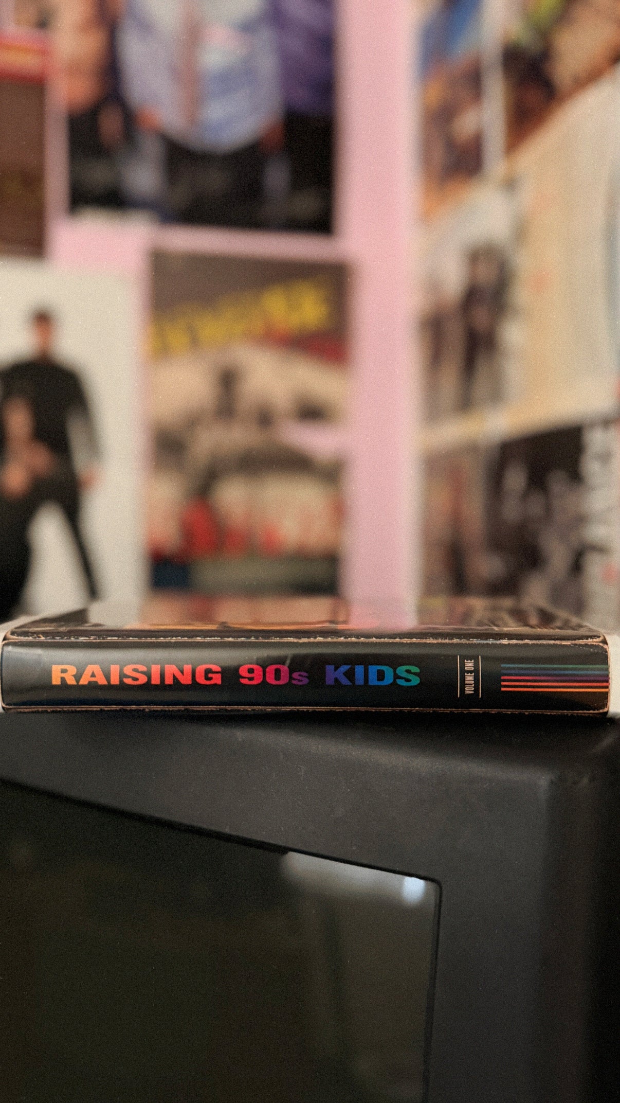 A VHS tape of the movie 'Raising 90's Kids' resting on top of a dark television set, with a blurred background of posters on a pink wall.