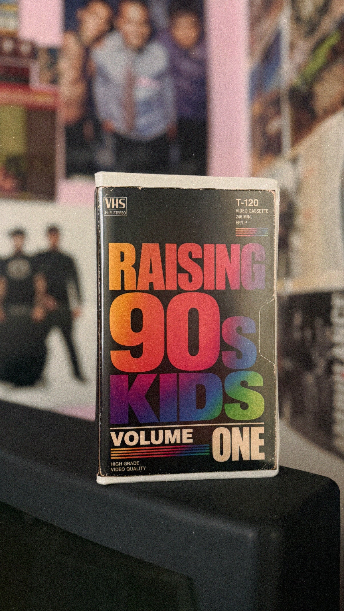 A VHS tape of the band 'Raising 90s Kids Volume One' standing upright on a black surface, with a blurred background of posters and photos.