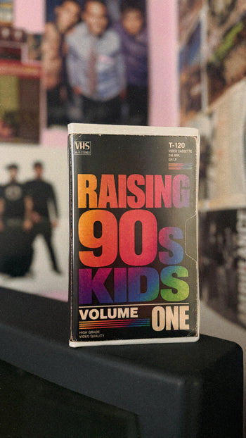 A VHS tape of the band 'Raising 90s Kids Volume One' standing upright on a black surface, with a blurred background of posters and photos.