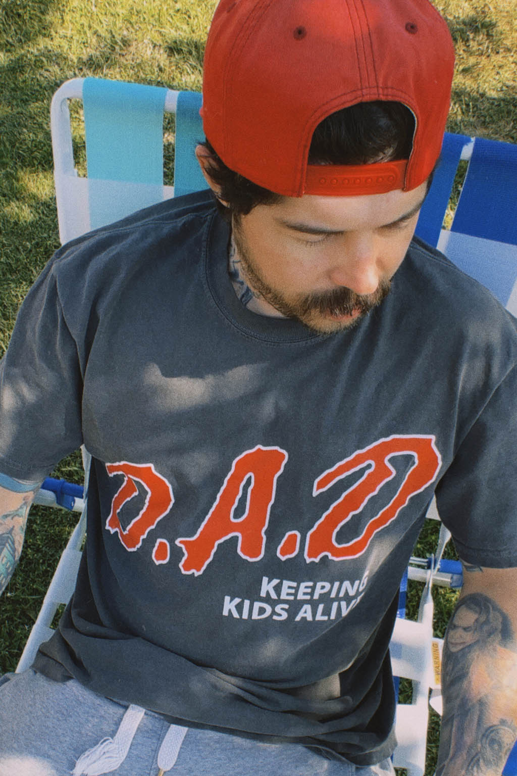 Man seated in a striped lawn chair on grass, wearing a backward red snapback cap and a faded black "D.A.D. Keeping Kids Alive" graphic tee with red lettering.