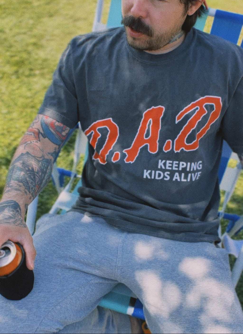 Tattooed man with a mustache relaxing in a striped lawn chair, holding a canned drink in a koozie, wearing a backward red snapback and a faded black "D.A.D. Keeping Kids Alive" graphic tee with gray sweatpants.