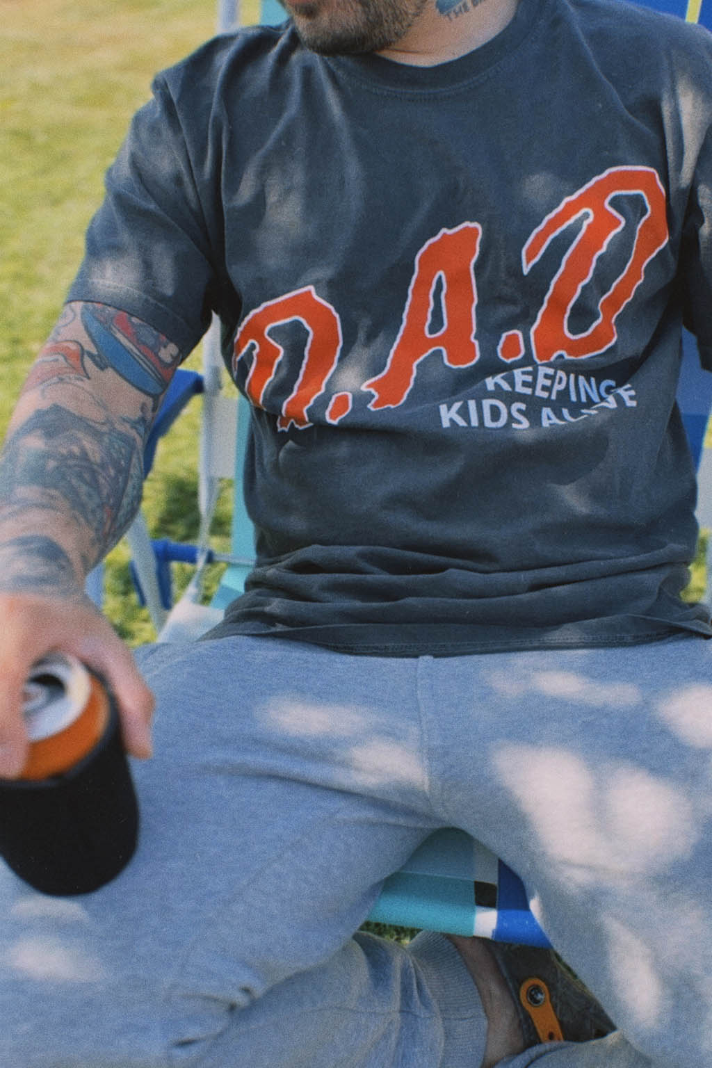Close-up of a man in a striped lawn chair holding a canned drink in a koozie, wearing the faded black "D.A.D. Keeping Kids Alive" graphic tee with red lettering and gray sweatpants.