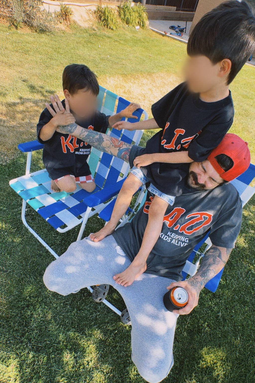 Father and two young sons in a sunny backyard wearing matching graphic tees from the "Keeping Kids Alive" family collection — dad in the "D.A.D." tee and the boys in coordinating "K.I.D." designs.