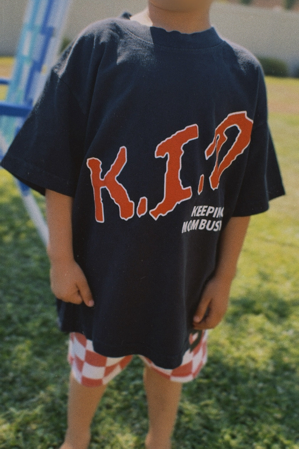 Child wearing a black t-shirt with 'K.I.D. KEEPING IT DUMB' text outdoors.