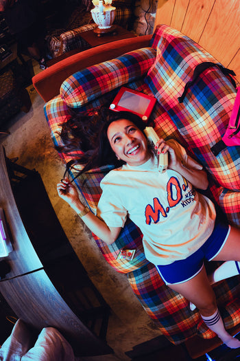 Smiling woman lying on a plaid couch holding a vintage cordless phone, wearing a cream "M.O.M." graphic tee and blue retro athletic shorts, with an Etch A Sketch beside her.