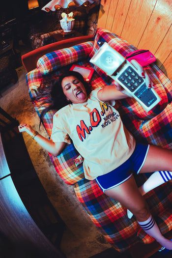Woman lying on a plaid couch with tongue out in playful exhaustion, holding out a vintage cordless phone, wearing the cream "M.O.M. Keeping Kids Alive" graphic tee and blue retro athletic shorts.