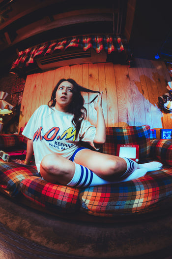 Woman seated on a plaid couch twirling her hair with a worried expression, wearing the cream "M.O.M. Keeping Kids Alive" graphic tee, blue retro athletic shorts, and white knee-high striped tube socks.