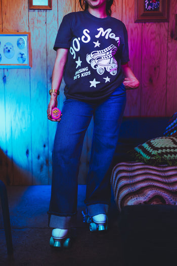 Person wearing a '90s Mom' t-shirt in a room with wooden walls and a bed.