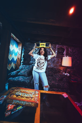 Person holding a skateboard above their head in a dimly lit room with a Jumanji game box on a table.