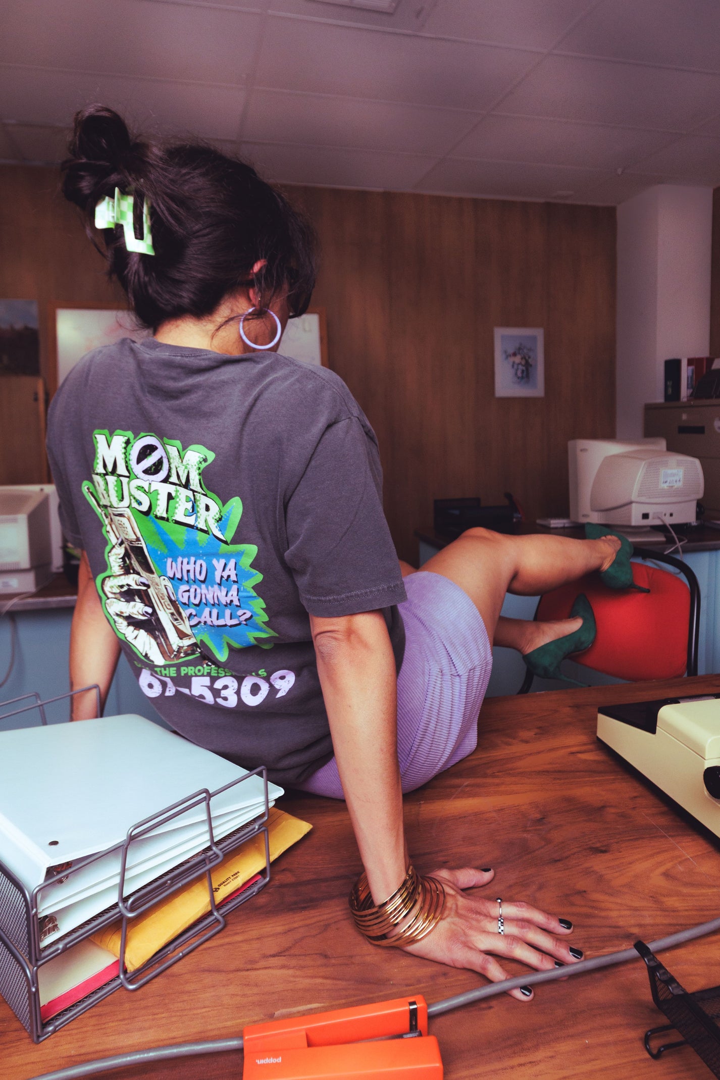 A woman seen from behind wearing a dark gray graphic t-shirt with a green and white 'Mom Operator' design, sitting at a wooden desk in an office environment.