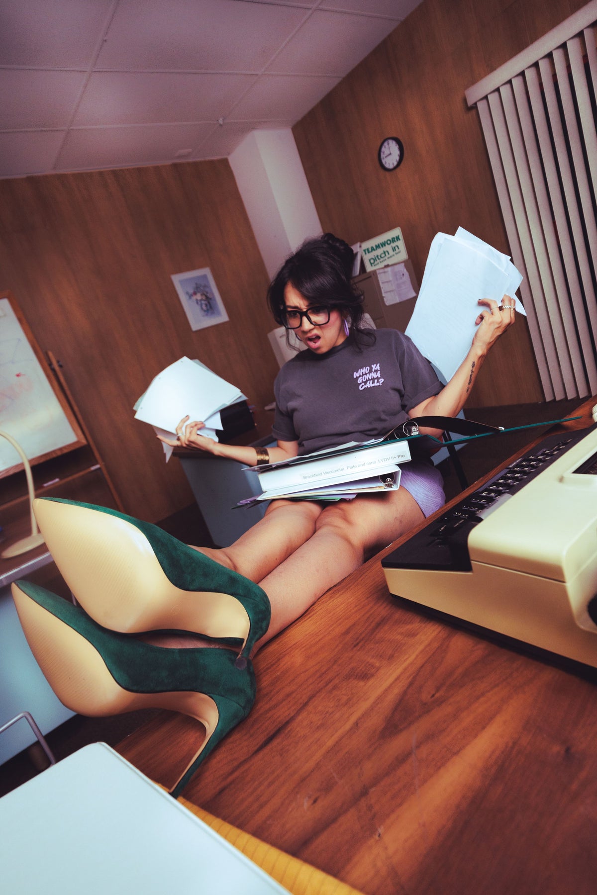 A woman in an office setting with wood-paneled walls sits on a desk, holding stacks of papers. She is wearing a dark gray t-shirt, light purple shorts, and distinctive dark green high heels with beige soles.