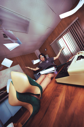A woman in an office setting with a typewriter and papers, wearing a very large, exaggerated green and beige high-heeled shoe on her foot, which dominates the foreground of the tilted photo.