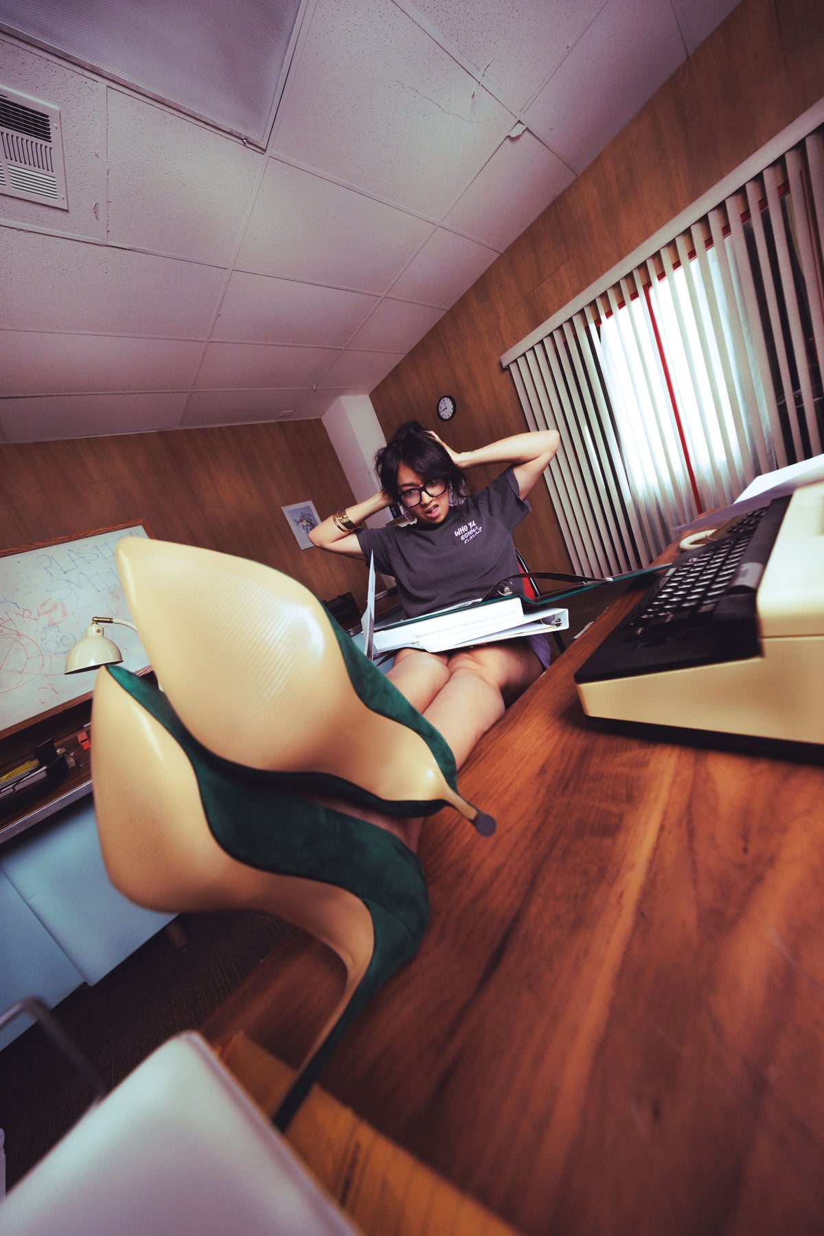 A person sitting at a desk in an office-like room, wearing oversized novelty high heels with a large beige front and dark green heels extending behind them. A typewriter, blinds, and wood-paneled walls are visible in the scene.