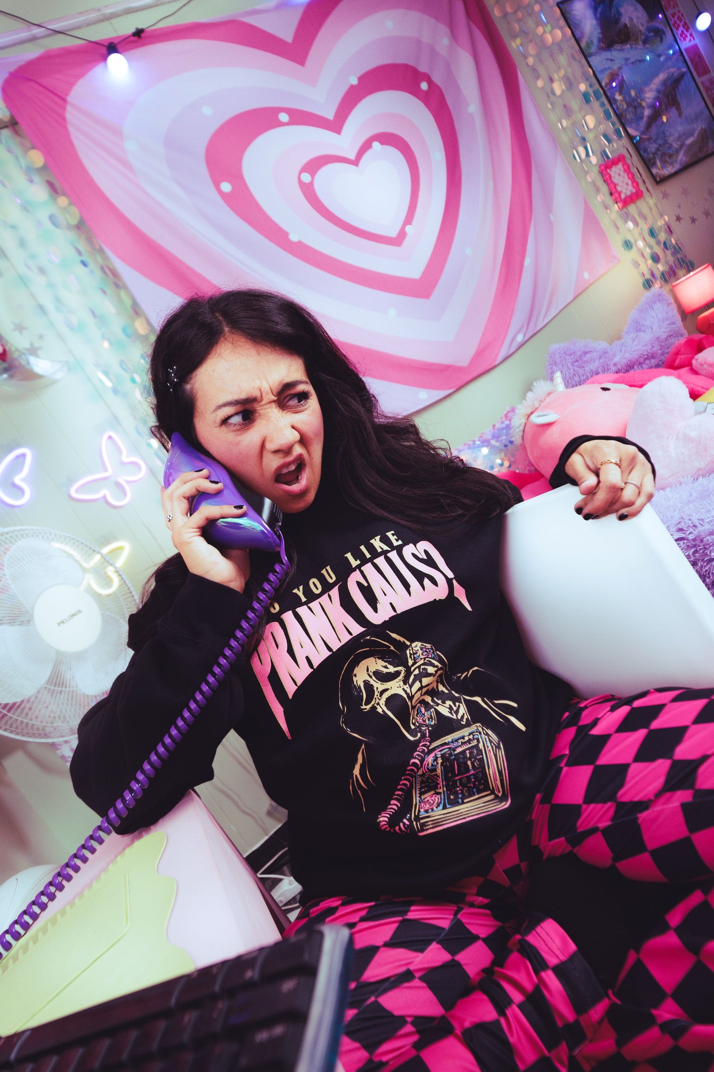 A woman in a pink and black checkered pajama pants and a black graphic sweatshirt holding a purple phone receiver, sitting in a room decorated with pink heart graphics.