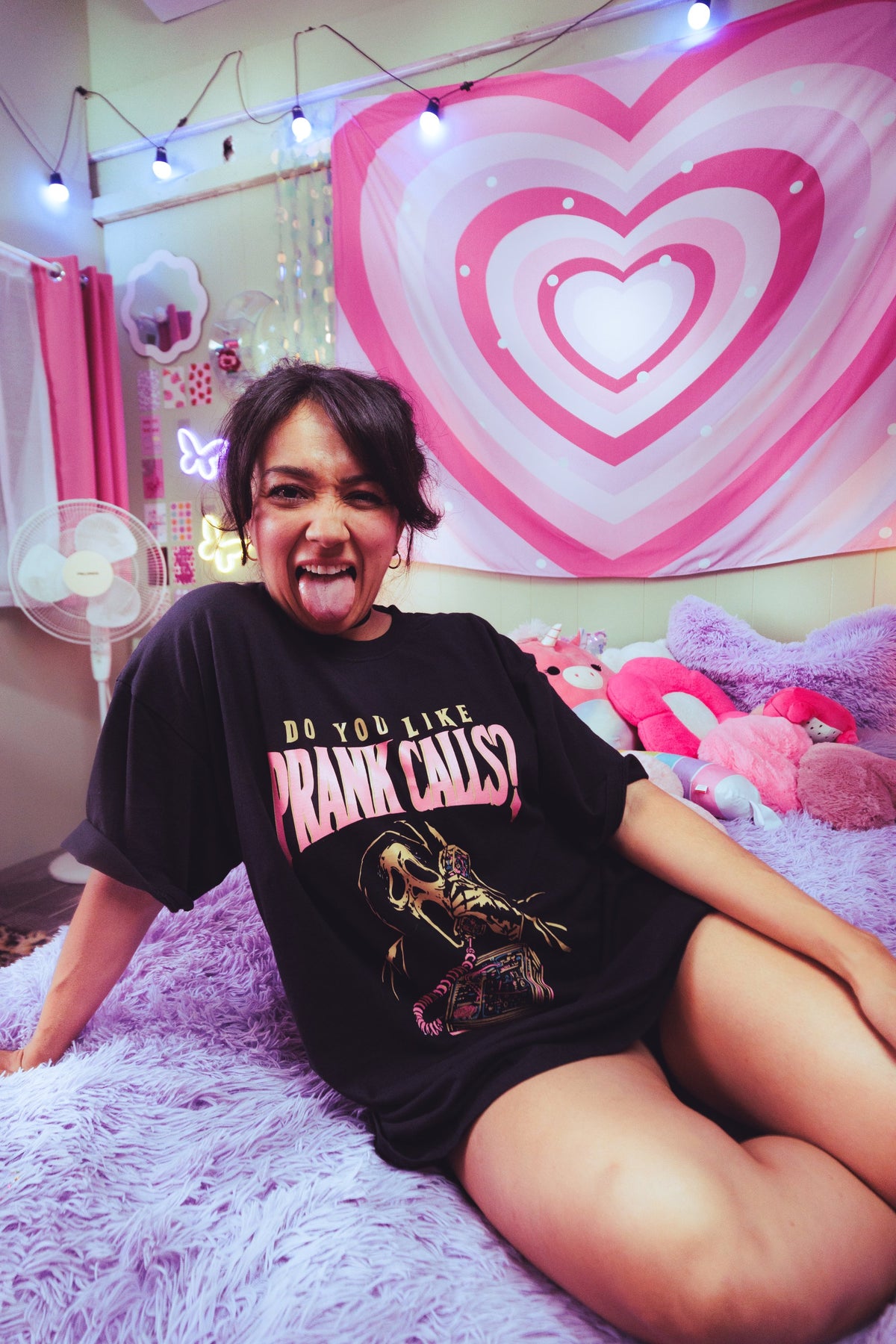 A person sitting on a fluffy lavender bed wearing an oversized black graphic T-shirt with the text 'DO YOU LIKE PRANK CALLS?' and a horror character print. The background features a large pink and white heart tapestry, string lights, and plush toys.