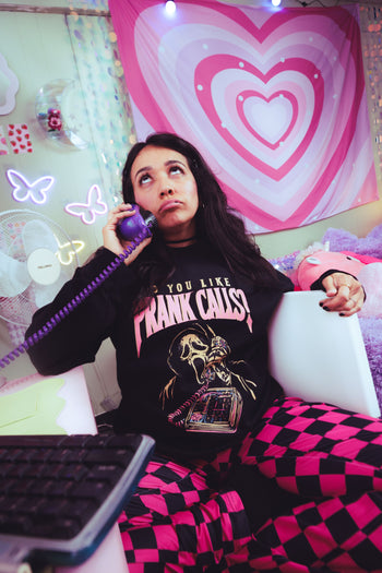 A woman sitting in a bedroom-like setting, holding a purple corded telephone receiver to her ear. She wears a black graphic T-shirt with pink text reading 'YOU YOU LIKE PRANK CALLS' and bright pink-and-black checkered pants.