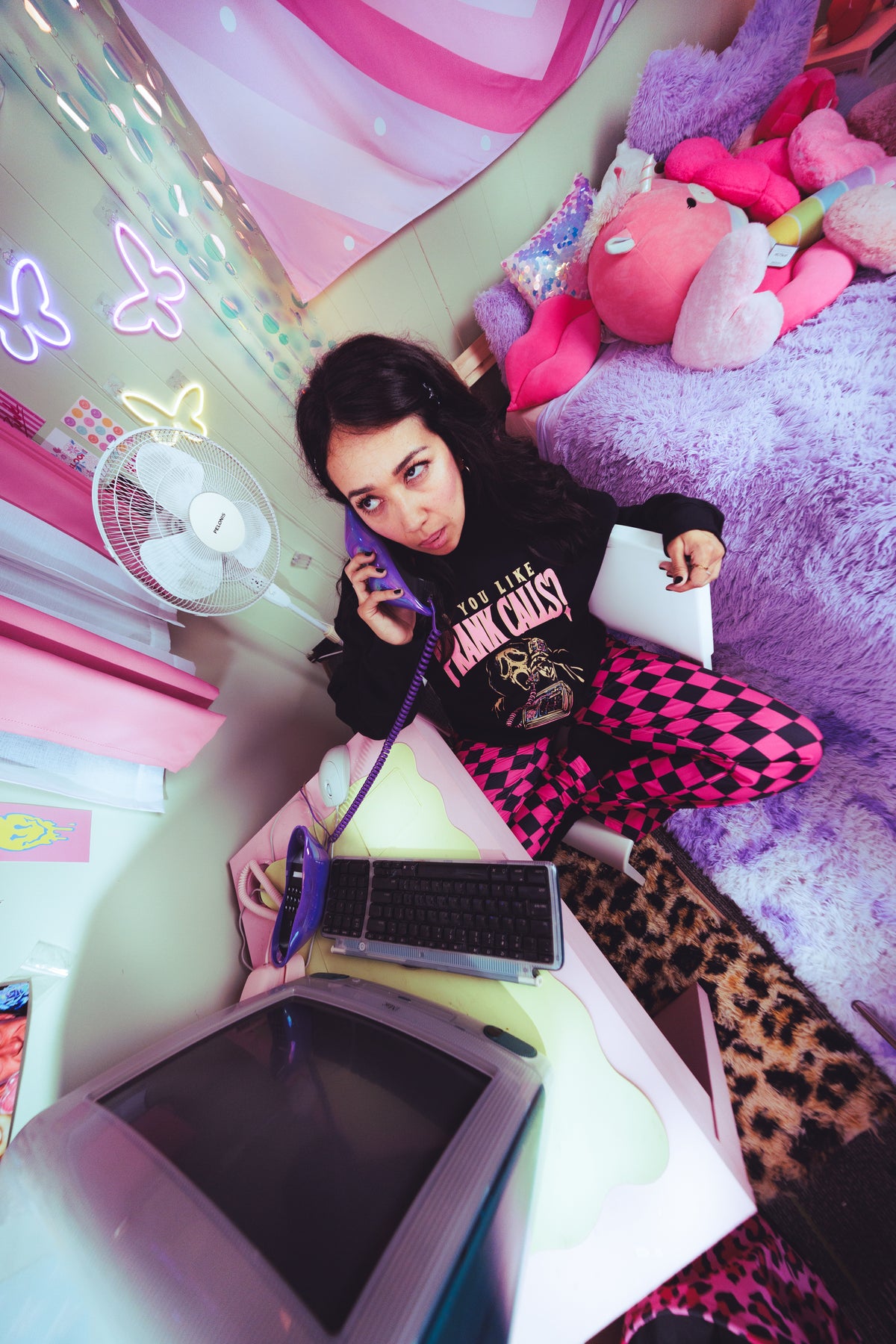 A young woman sits on a bed in a brightly lit bedroom, holding a purple corded telephone receiver and looking at a white tablet. She wears a black graphic sweatshirt and pink and black checkered leggings.