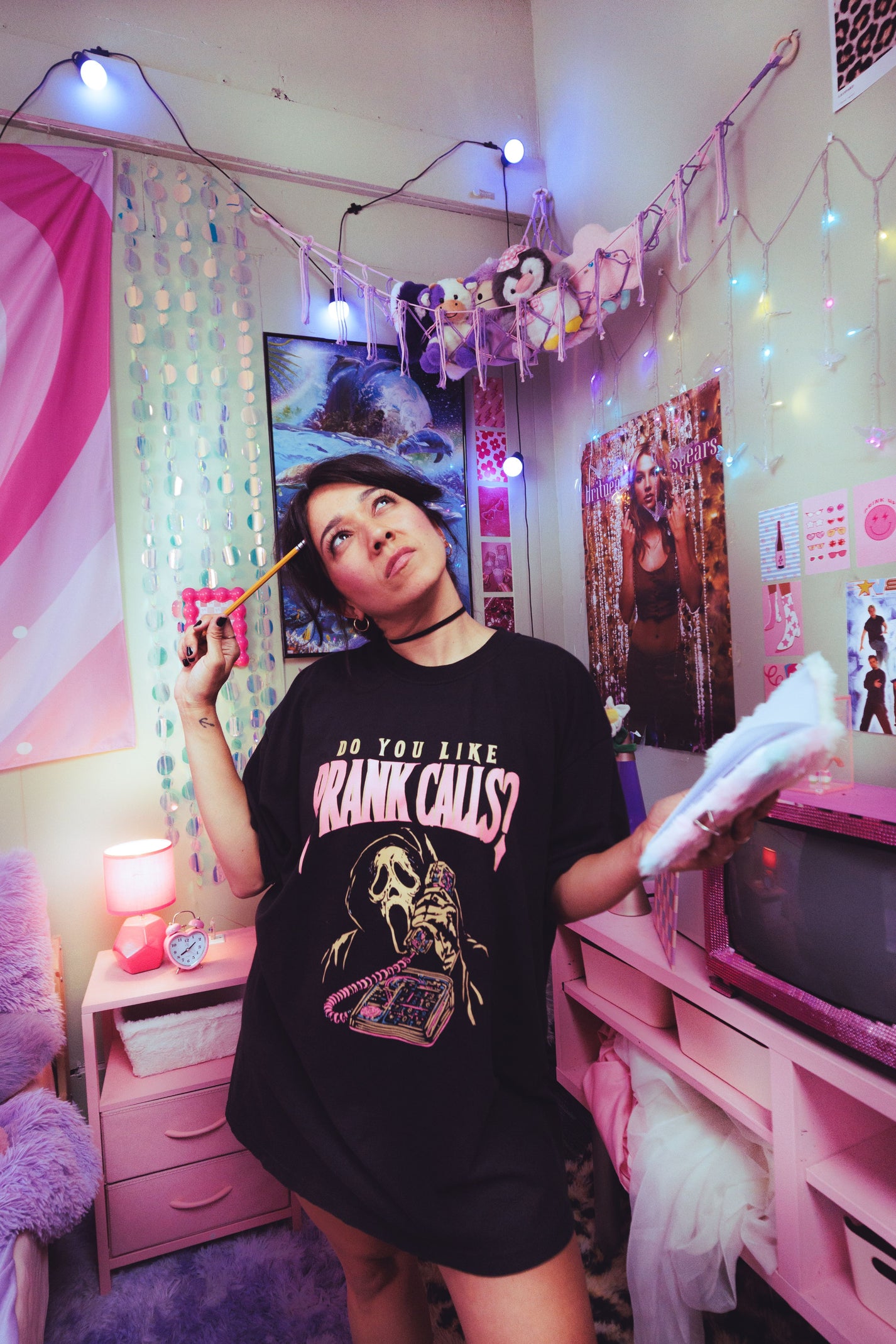 A person standing in a pink bedroom wearing an oversized black graphic T-shirt with the text 'DO YOU LIKE PRANK CALLS?' and a horror character print. The room has string lights, posters, a pink dresser, and a mirror.