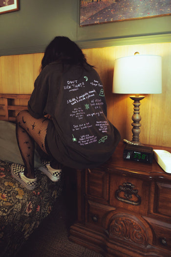 A person sitting on a bed in a bedroom, viewed from behind, wearing an oversized dark brown T-shirt with handwritten white text and graphics on the back. The room has a wooden nightstand with a lit lamp, and the person is also wearing fishnet tights and sneakers.