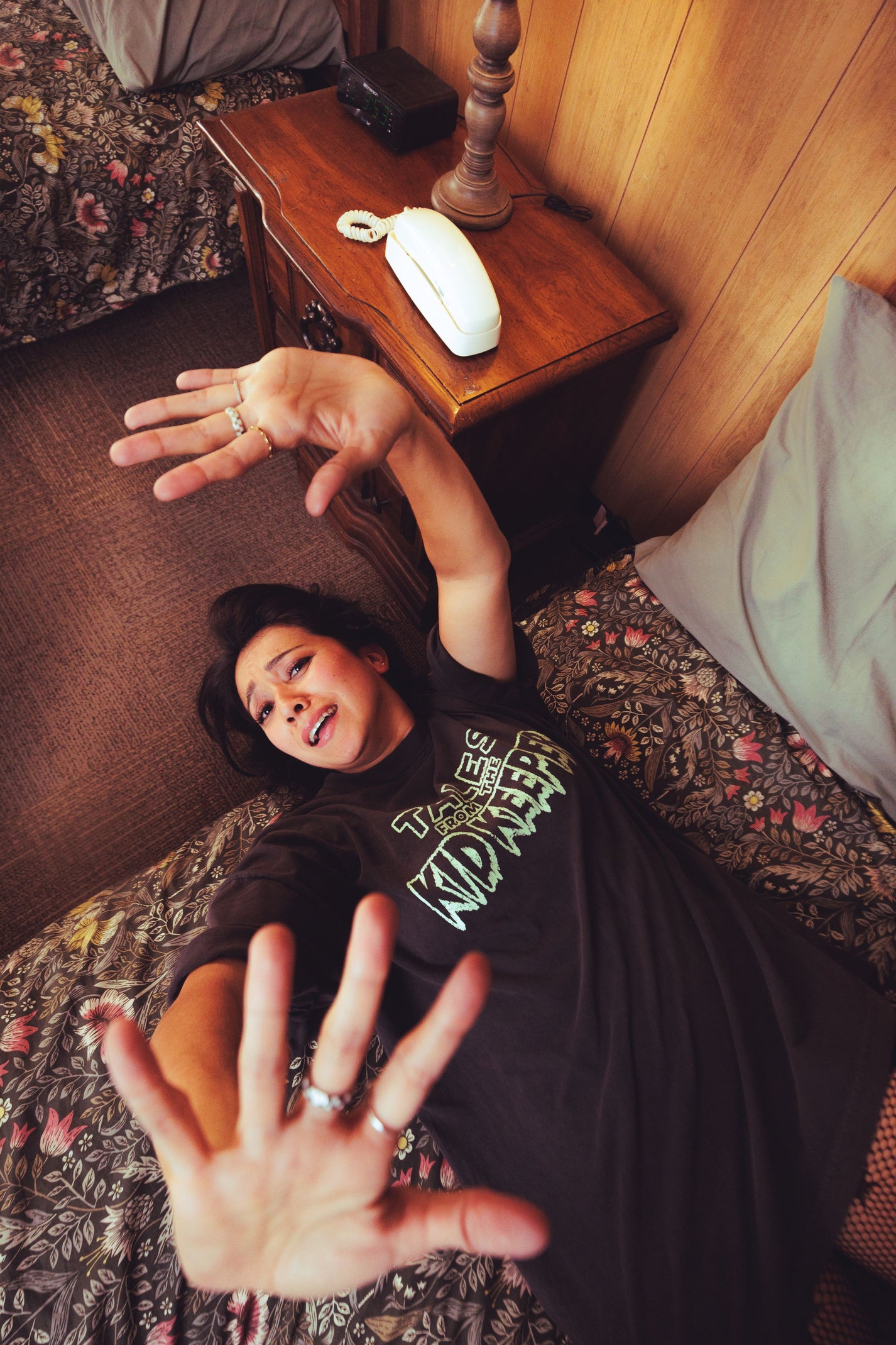 A woman lying on a bed in a bedroom, wearing a black graphic t-shirt with green and white text that reads 'Tales of the Kid Keeper'. She is reaching toward the camera with both hands.
