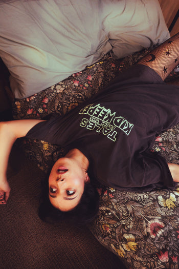 A person lying on a floral-patterned bed wearing a dark oversized t-shirt with 'TALES OF KID KEEPER' printed in white and green on the front, paired with fishnet stockings.