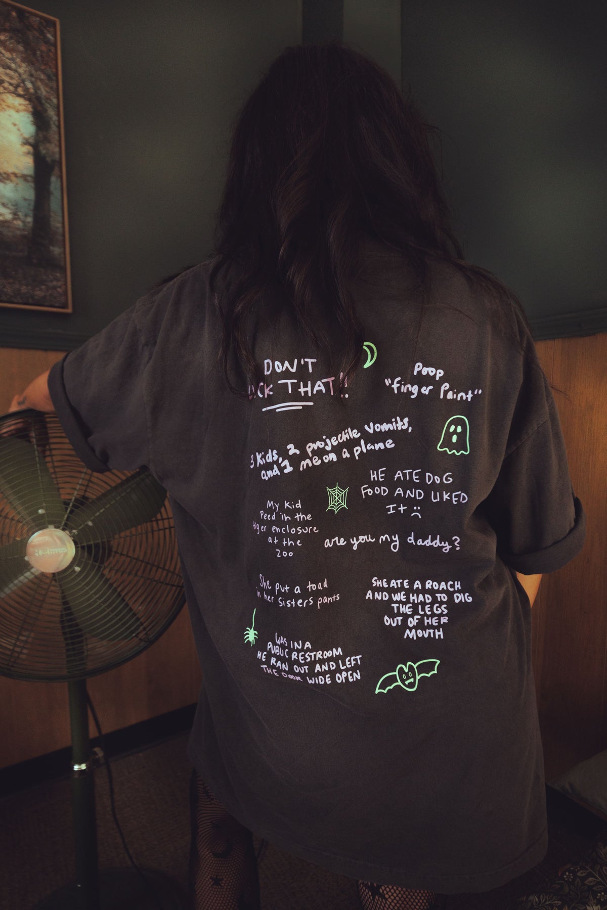 A person viewed from behind wearing an oversized dark gray T-shirt with handwritten white and neon green text and graphics on the back, standing indoors near a floor fan in a dim room.