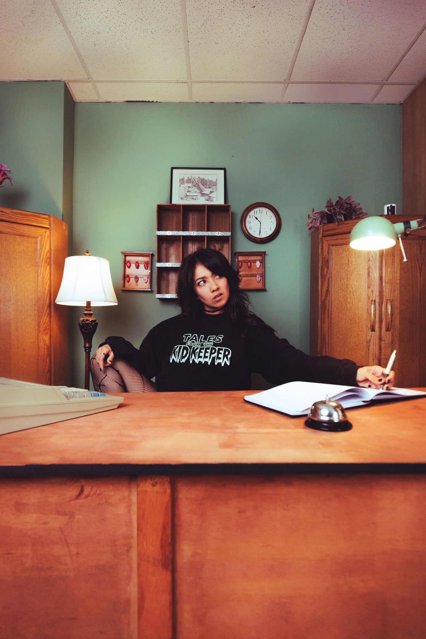 A woman sits behind a wooden reception desk in an office, wearing a black sweatshirt with white text that reads 'TALES OF THE KID KEEPER''. The room has green walls, wooden cabinets, a lamp, a clock, and a bell on the desk.