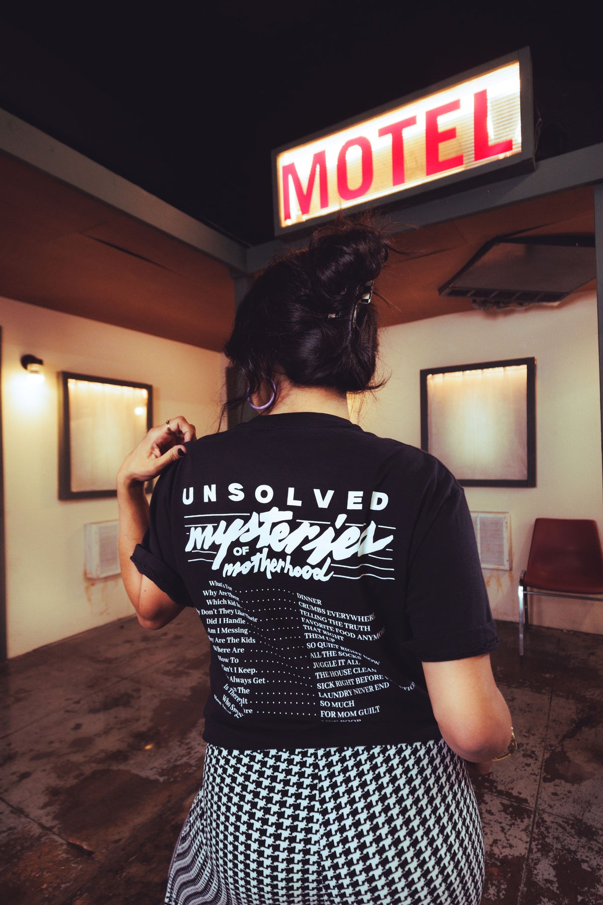 A person viewed from behind wearing a black graphic T-shirt with white text reading 'Unsolved Mysteries of Motherhood' and a black-and-white houndstooth skirt, standing in front of a glowing red-and-white 'MOTEL' sign in a dim interior.