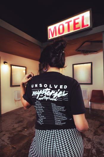 A person viewed from behind wearing a black graphic T-shirt with white text reading 'Unsolved Mysteries of Motherhood' and a black-and-white houndstooth skirt, standing in front of a glowing red-and-white 'MOTEL' sign in a dim interior.