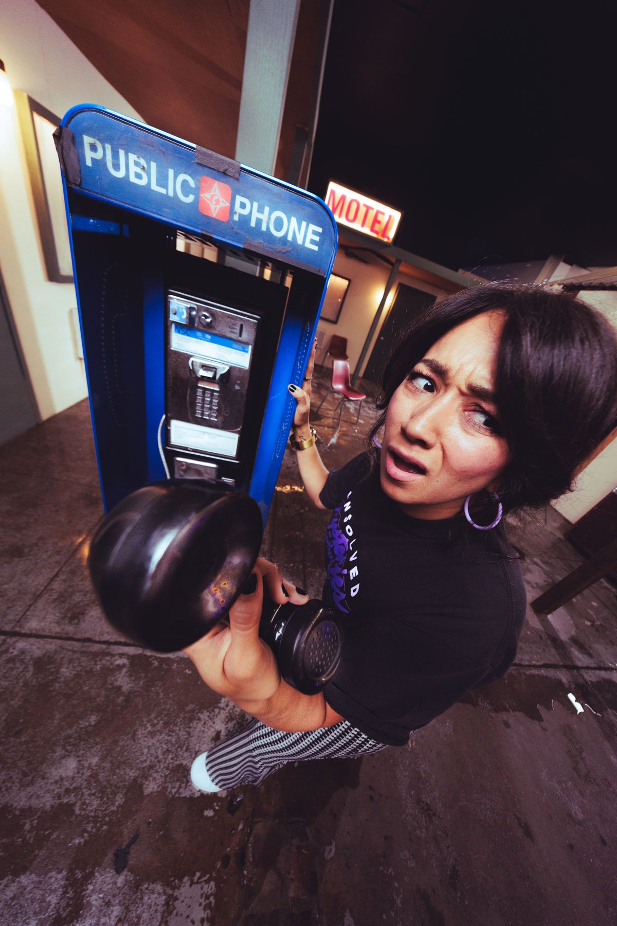 A woman in a black graphic t-shirt and striped pants poses with a black gas mask in front of a blue public phone booth at night.