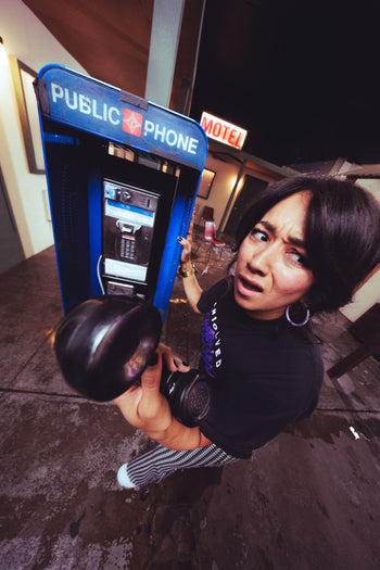 A woman in a black graphic t-shirt and striped pants poses with a black gas mask in front of a blue public phone booth at night.