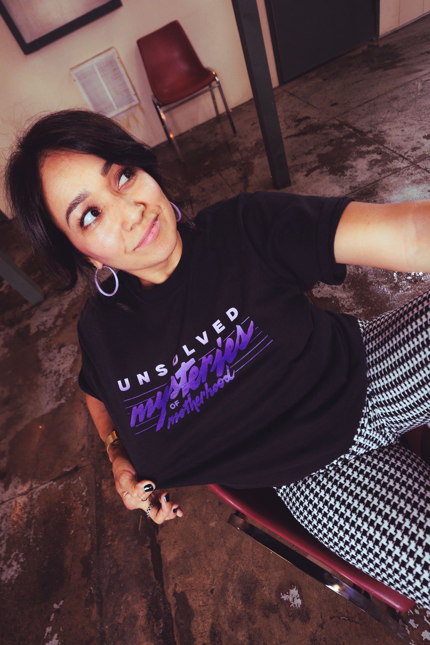 A woman wearing a black graphic t-shirt with 'Unsolved Mysteries of Motherhood' printed in purple and white, paired with black and white checkered pants, captured in a casual indoor setting.