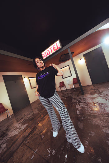 A woman posing in a motel lobby, wearing a black graphic t-shirt with purple text and black-and-white checkered wide-leg pants.